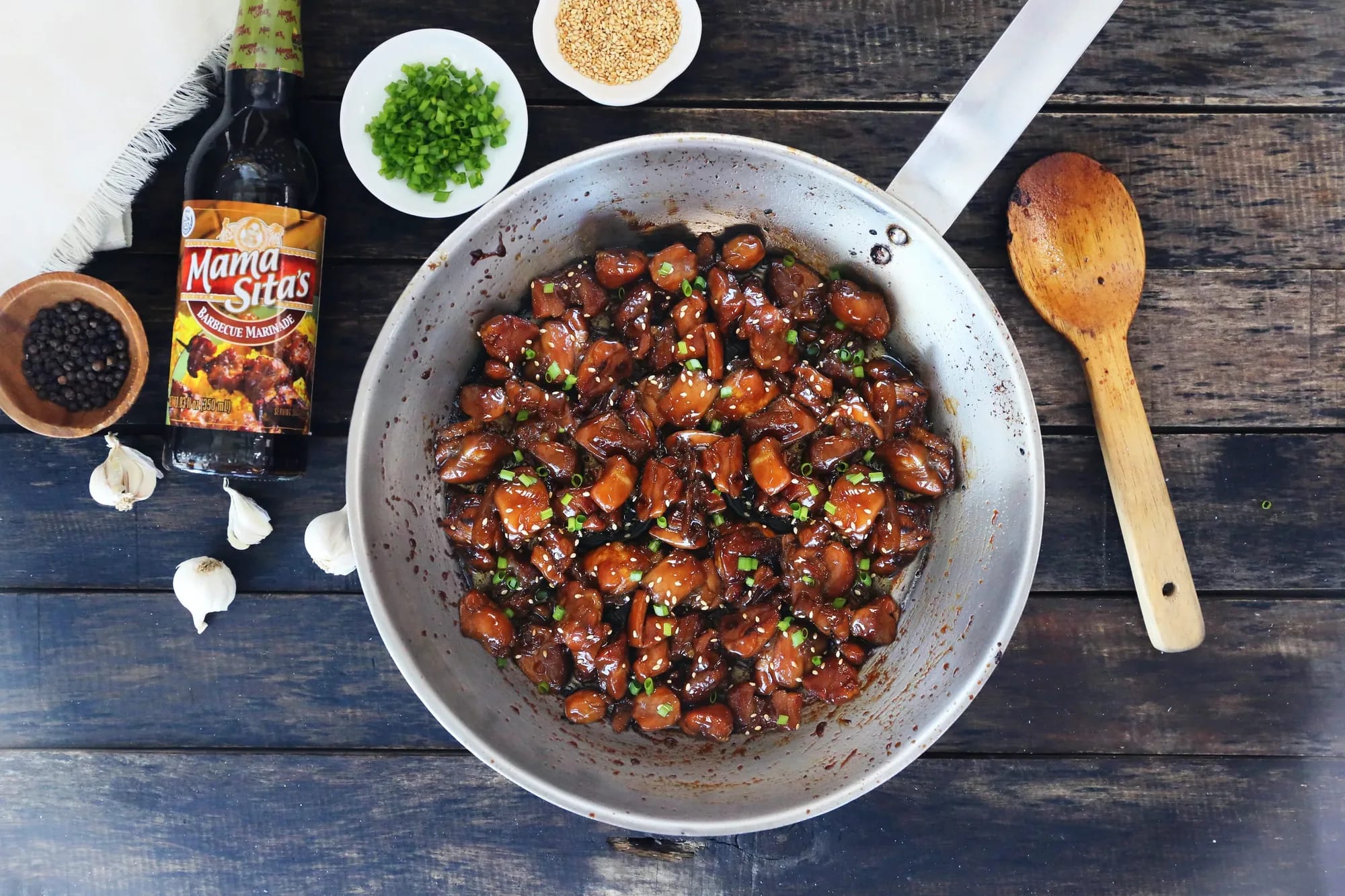Chicken barbecue glazing in a pan on a dark wooden table, the Mama Sita's Barbecue Marinade bottle beside it