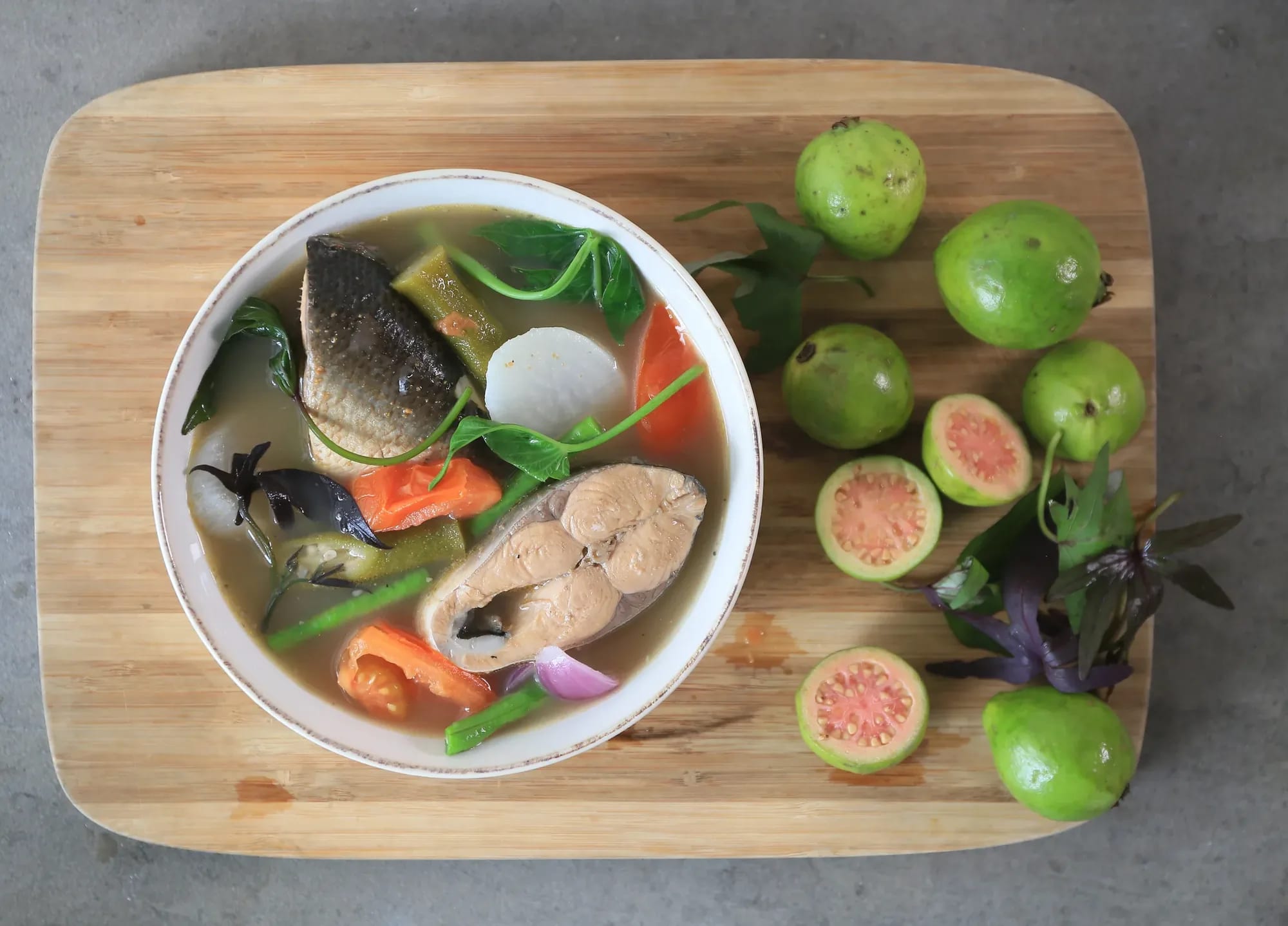 Sinigang na bangus in a white bowl on a wooden board, ringed with halved green guavas