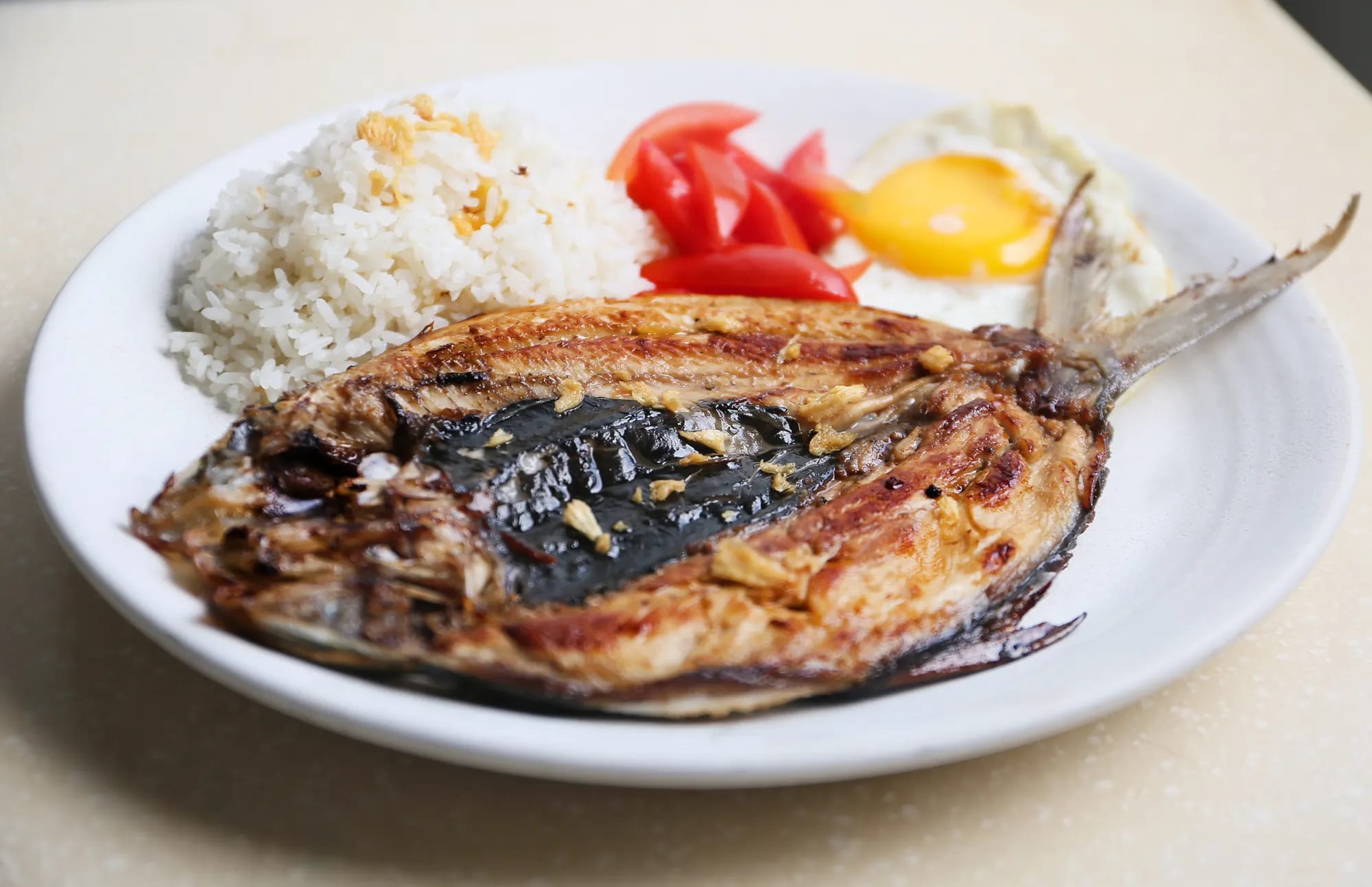 A butterflied grilled milkfish with garlic rice, sliced tomato and a fried egg on a white plate, on a cream speckled table