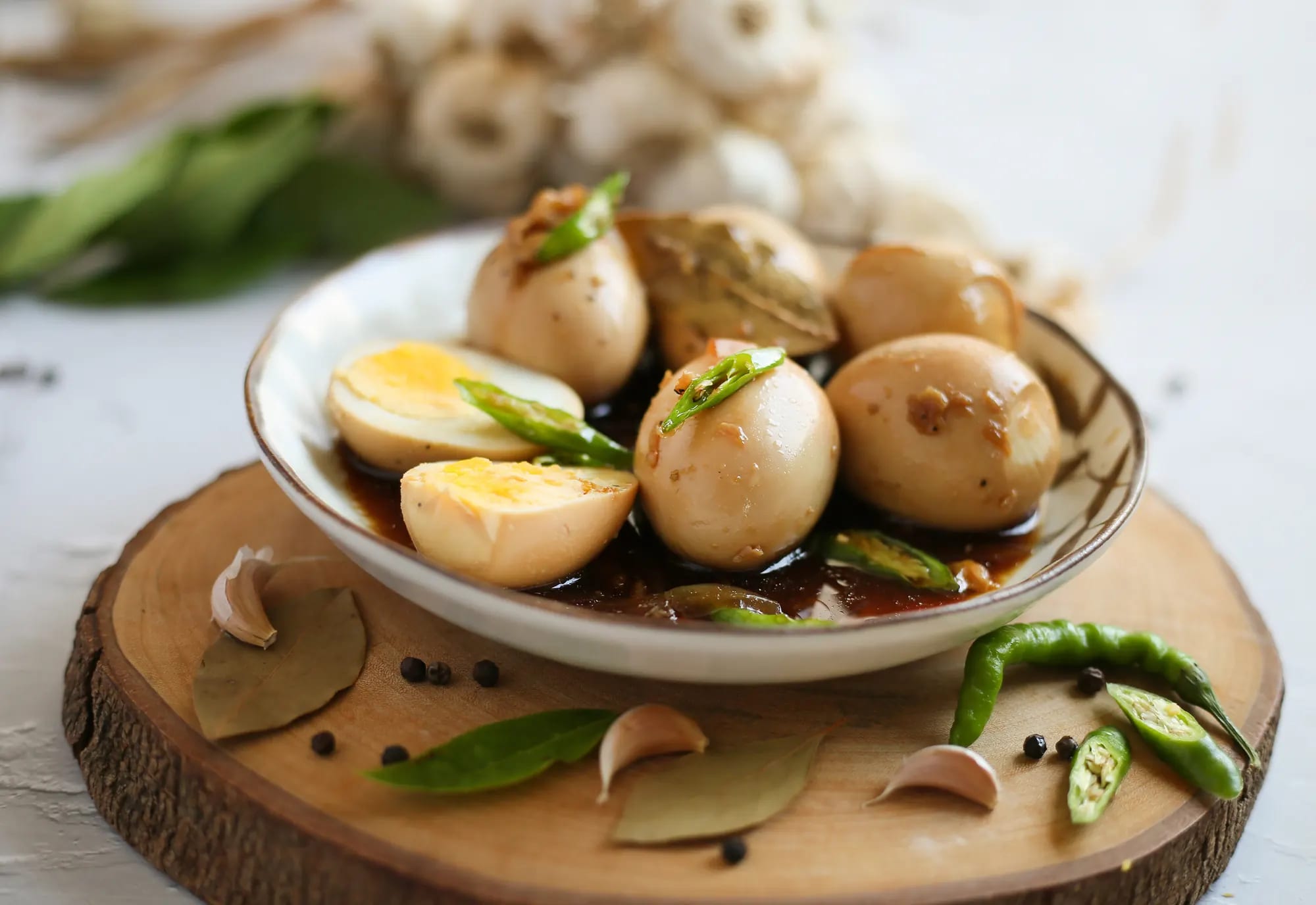 Whole and halved hard-boiled eggs in dark adobo sauce in a cream bowl, on a live-edge wooden round with bay leaves, garlic and green chilies