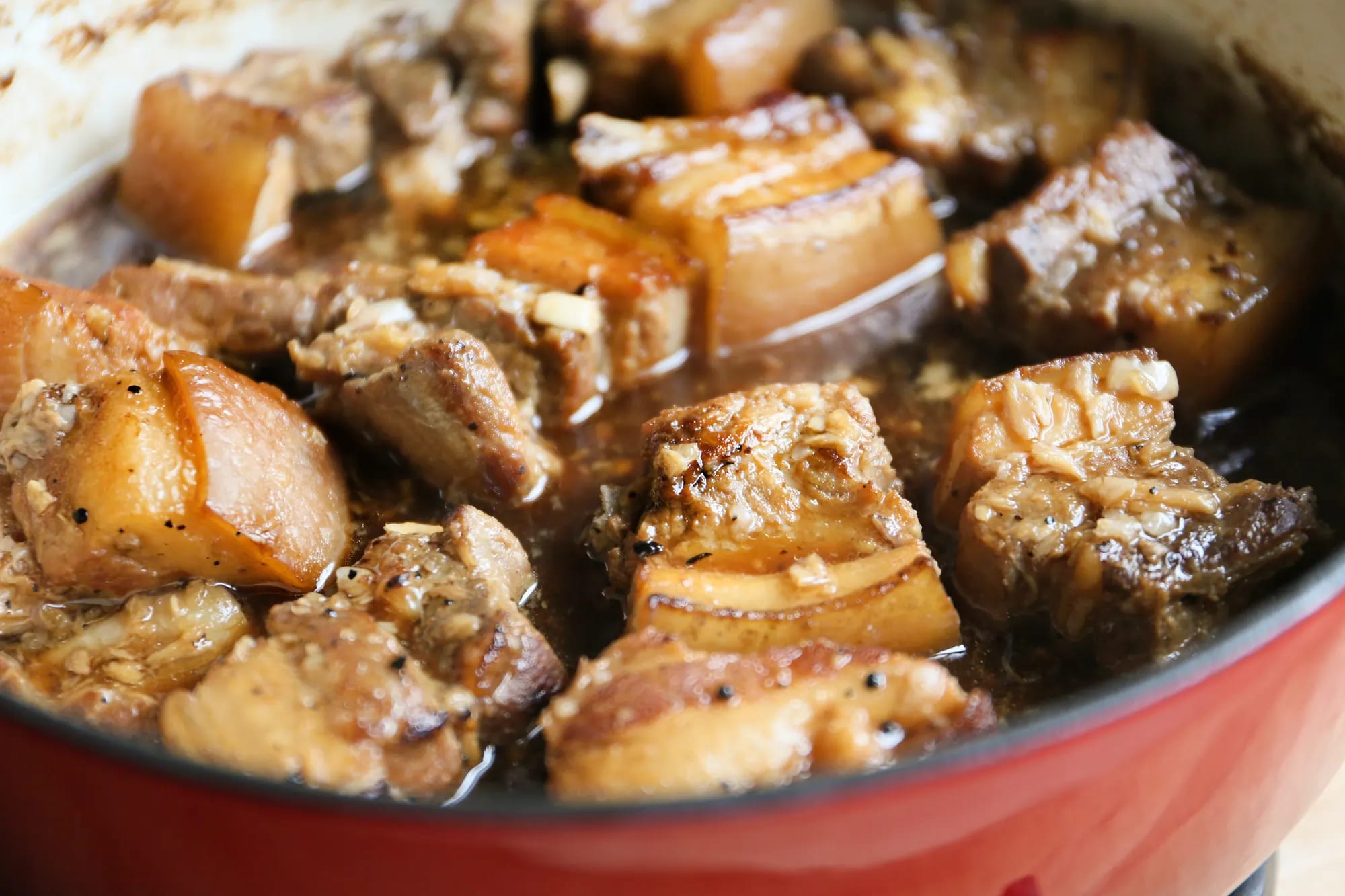 Cubes of braised pork belly with garlic and peppercorns in dark adobo sauce, filling a red enamelled pot, photographed from directly above