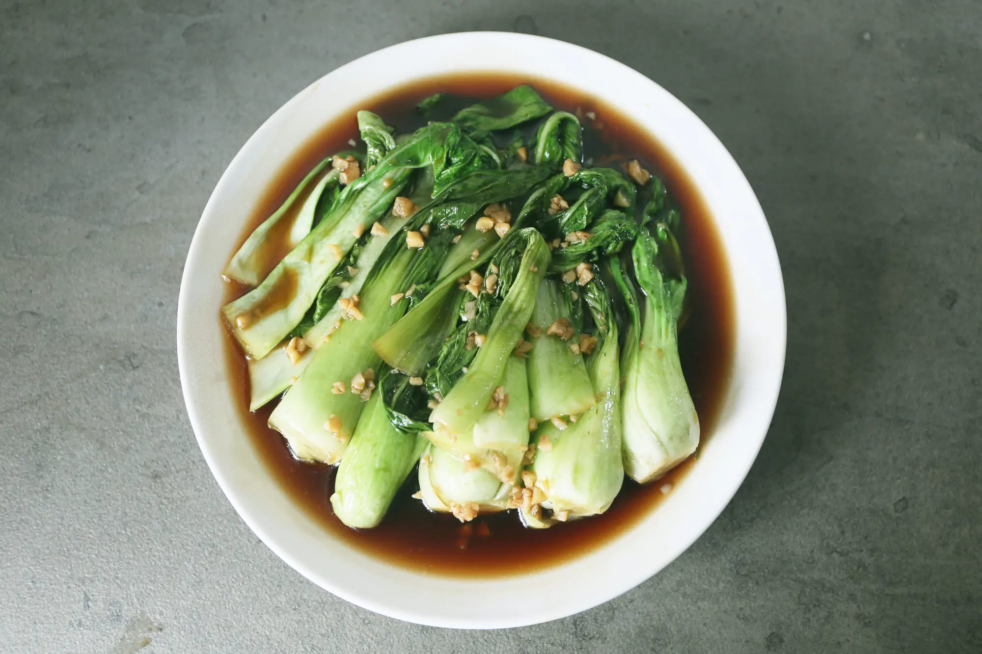 Halved heads of bok choy in a dark oyster-sauce glaze with fried garlic in a white bowl, photographed from above on grey concrete
