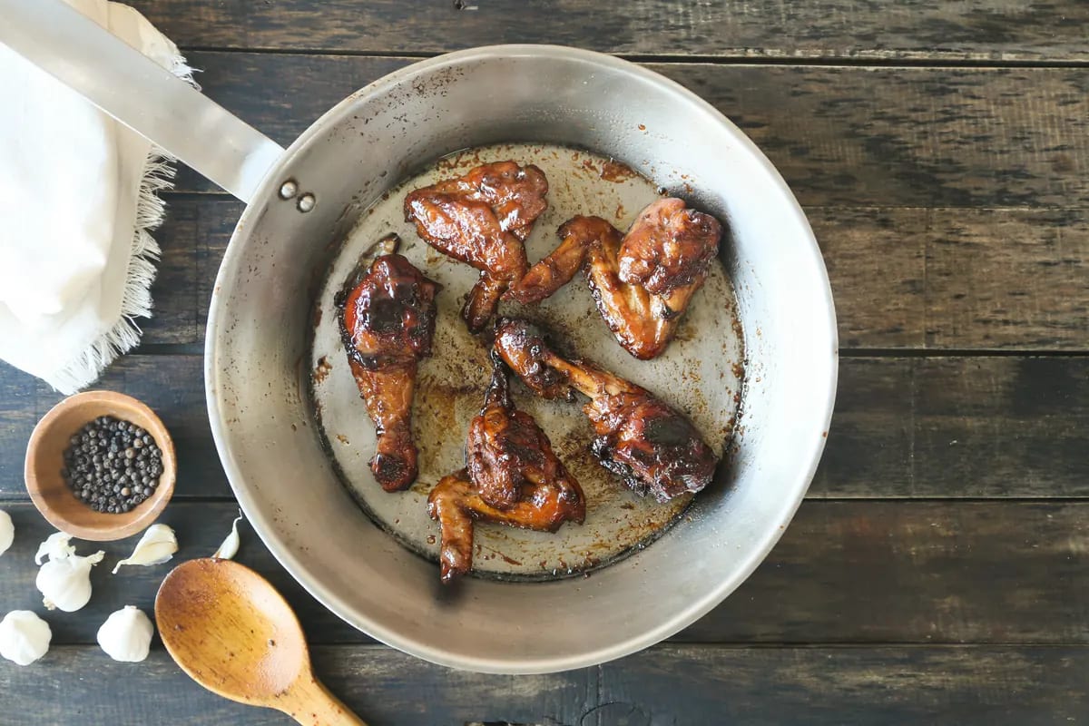 Six pieces of dark-glazed barbecued chicken in a wide metal pan, photographed from above on grey-blue weathered planks beside a small bowl of peppercorns, garlic cloves and a wooden spoon