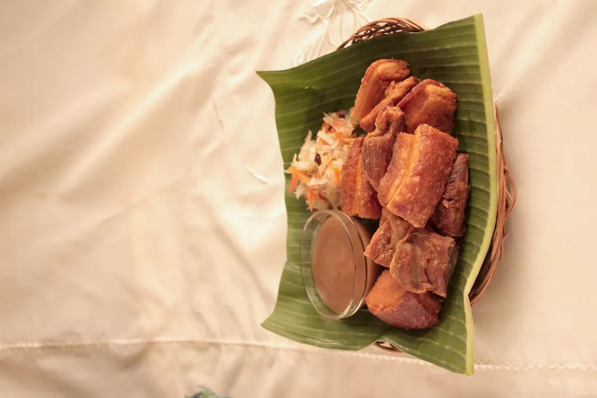 Six slabs of fried pork belly with crackled rind on a banana leaf in a round rattan basket, with pickled papaya relish and a small glass of brown sauce, on a cream cloth