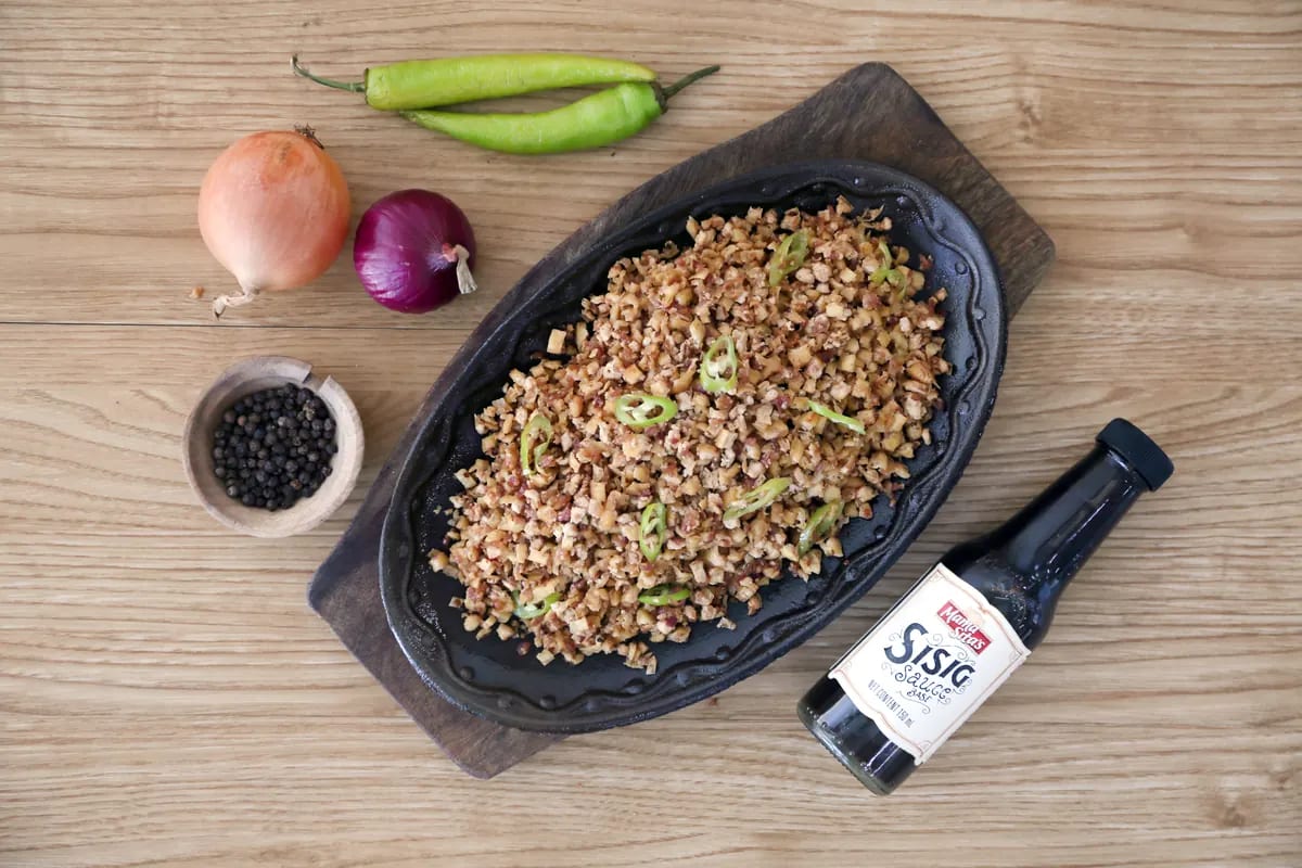 Chopped tofu sisig with sliced green chilies on a black cast-iron sizzling platter, photographed from above on pale wood beside onions, peppercorns and a bottle of Mama Sita's Sisig Sauce Base