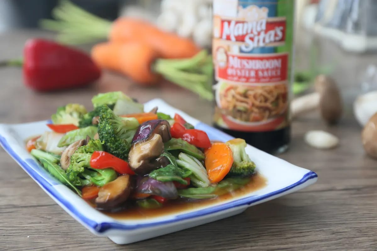 Stir-fried broccoli, bell pepper, carrot, mushrooms and beans in brown sauce on a blue-rimmed white plate, with a bottle of Mama Sita's Mushroom Oyster Sauce standing behind
