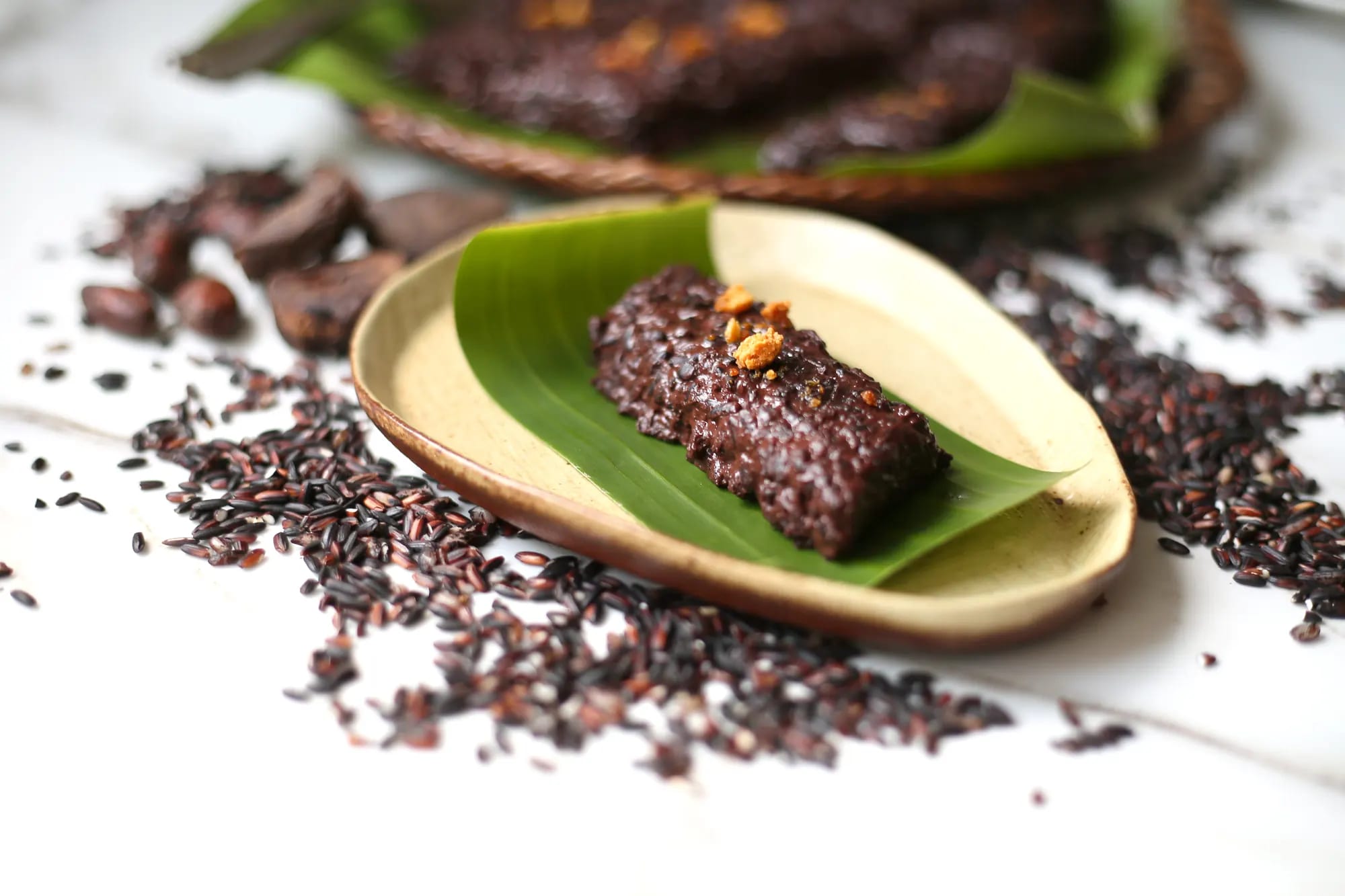 A bar of black glutinous rice cake topped with golden coconut curds, on a banana leaf in a shallow wooden dish, with raw black rice grains scattered on a white table