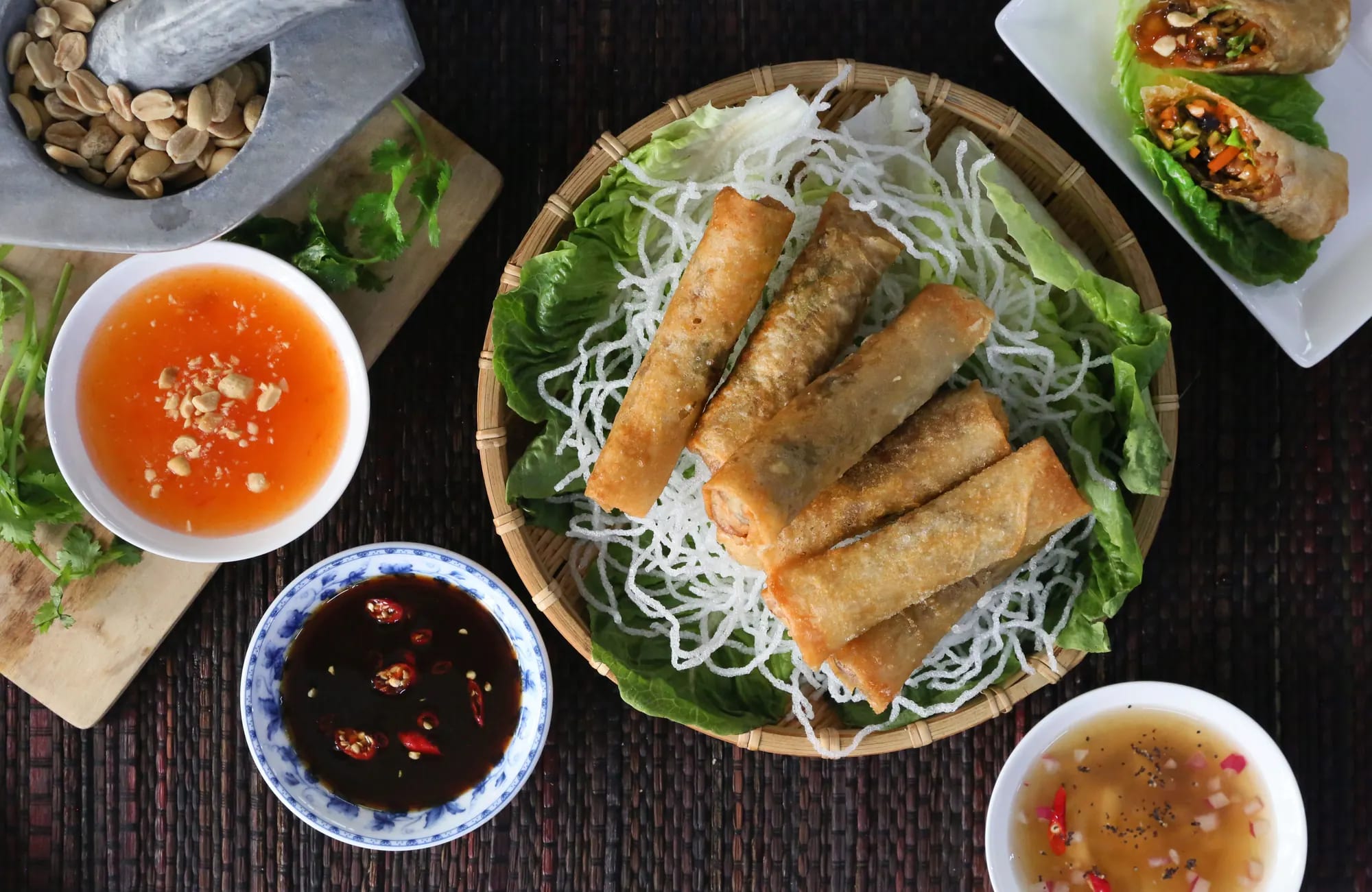 Six fried spring rolls on crisp rice noodles and lettuce in a round bamboo tray, photographed from above on a dark woven mat with three bowls of dipping sauce and a mortar of peanuts around it