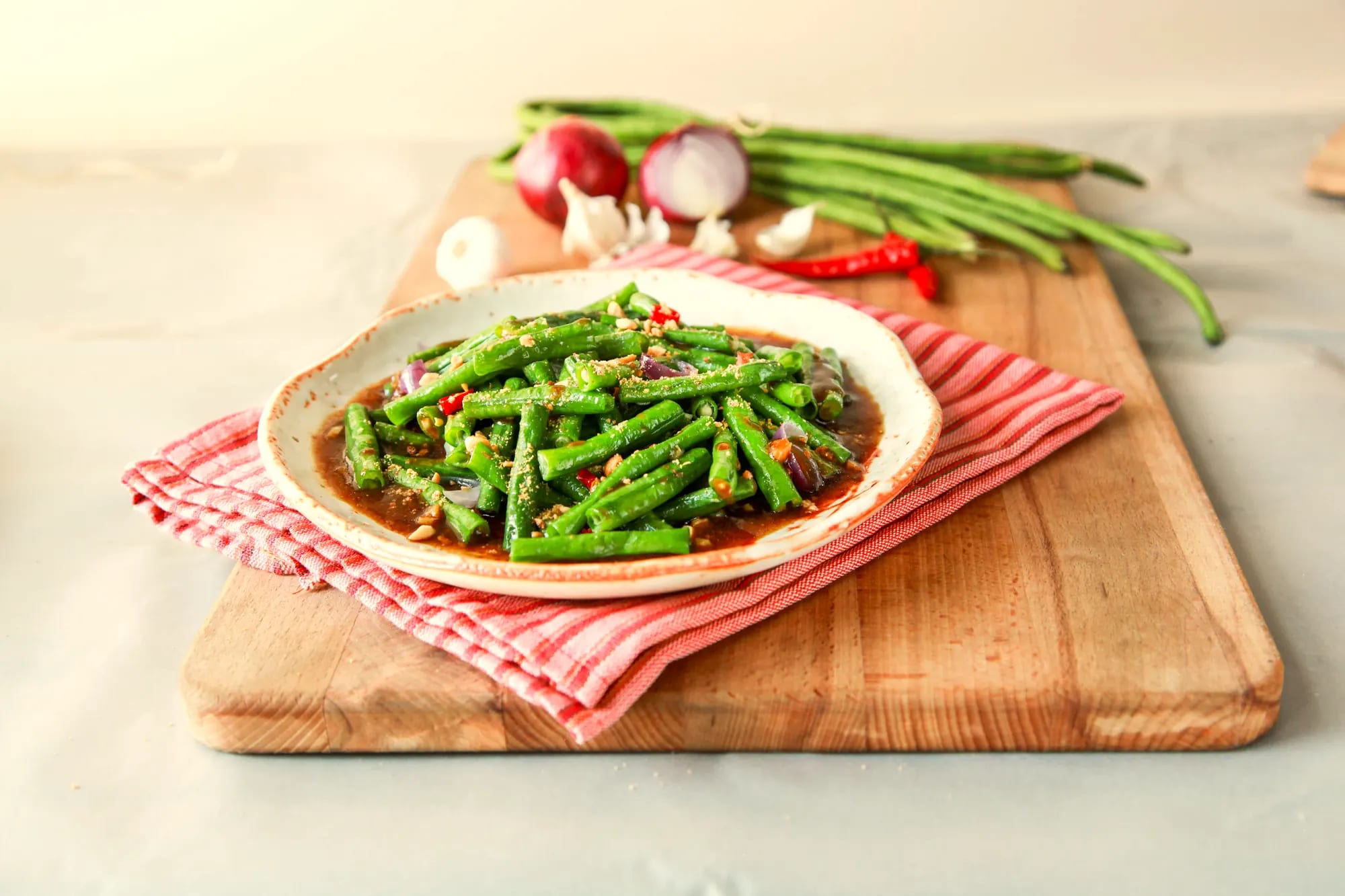 Sautéed yardlong beans with red onion and chili in brown sauce on a cream stoneware plate, on a red-and-white striped cloth over a wooden board, with onion, garlic and long beans behind