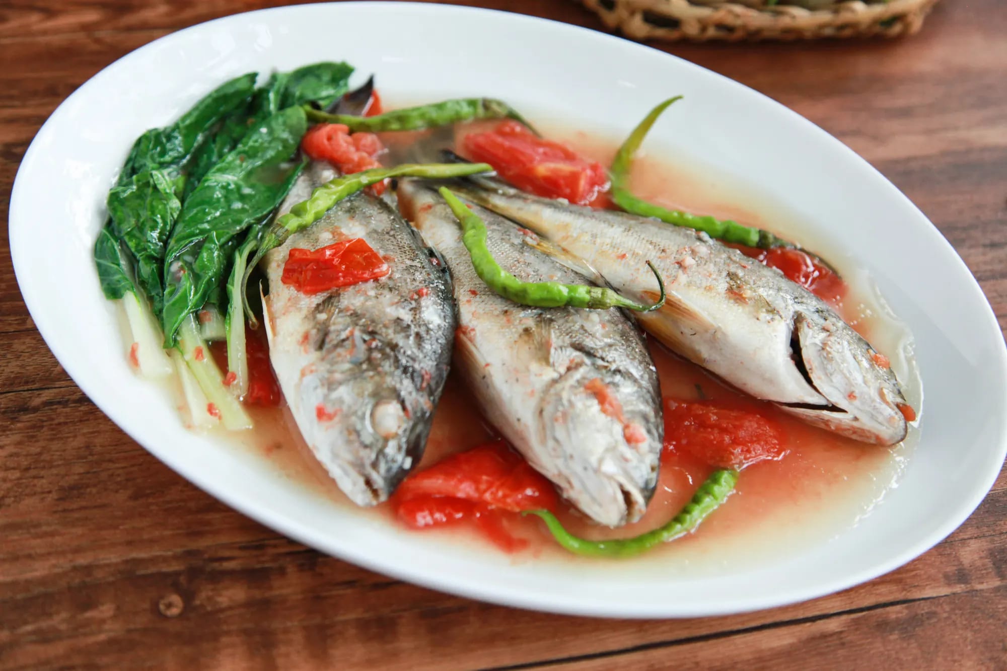 Three whole small fish in a pale sour broth with tomato, green chilies and mustard greens on a large white oval platter, photographed from above on a wooden table