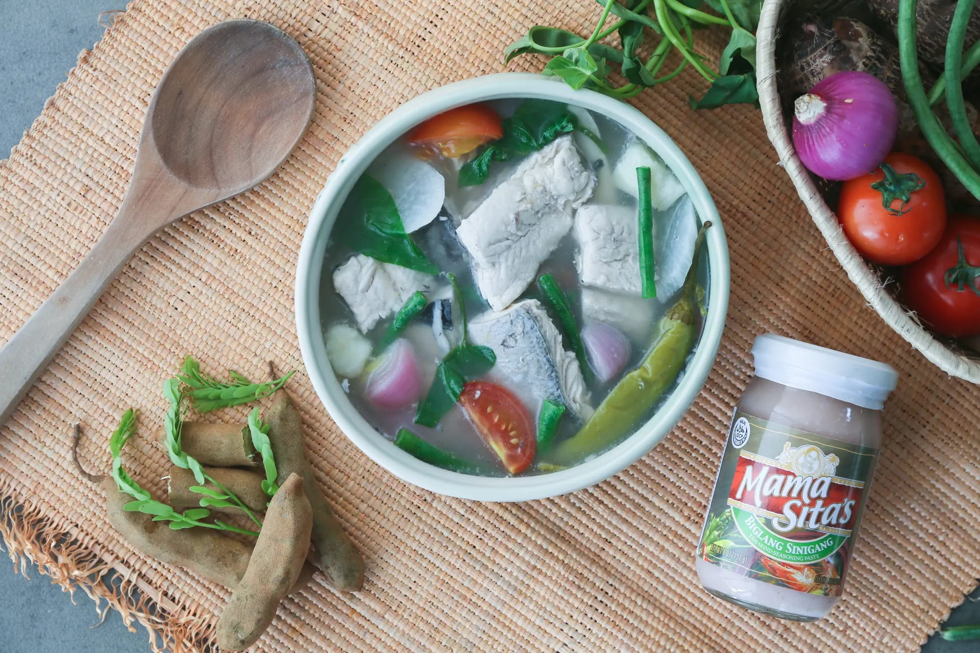 Milkfish steaks in a clear sour broth with green chilies, yardlong beans, tomato and radish in a pale bowl, photographed from above on a woven mat beside a wooden spoon, tamarind pods and a jar of Mama Sita's Biglang Sinigang