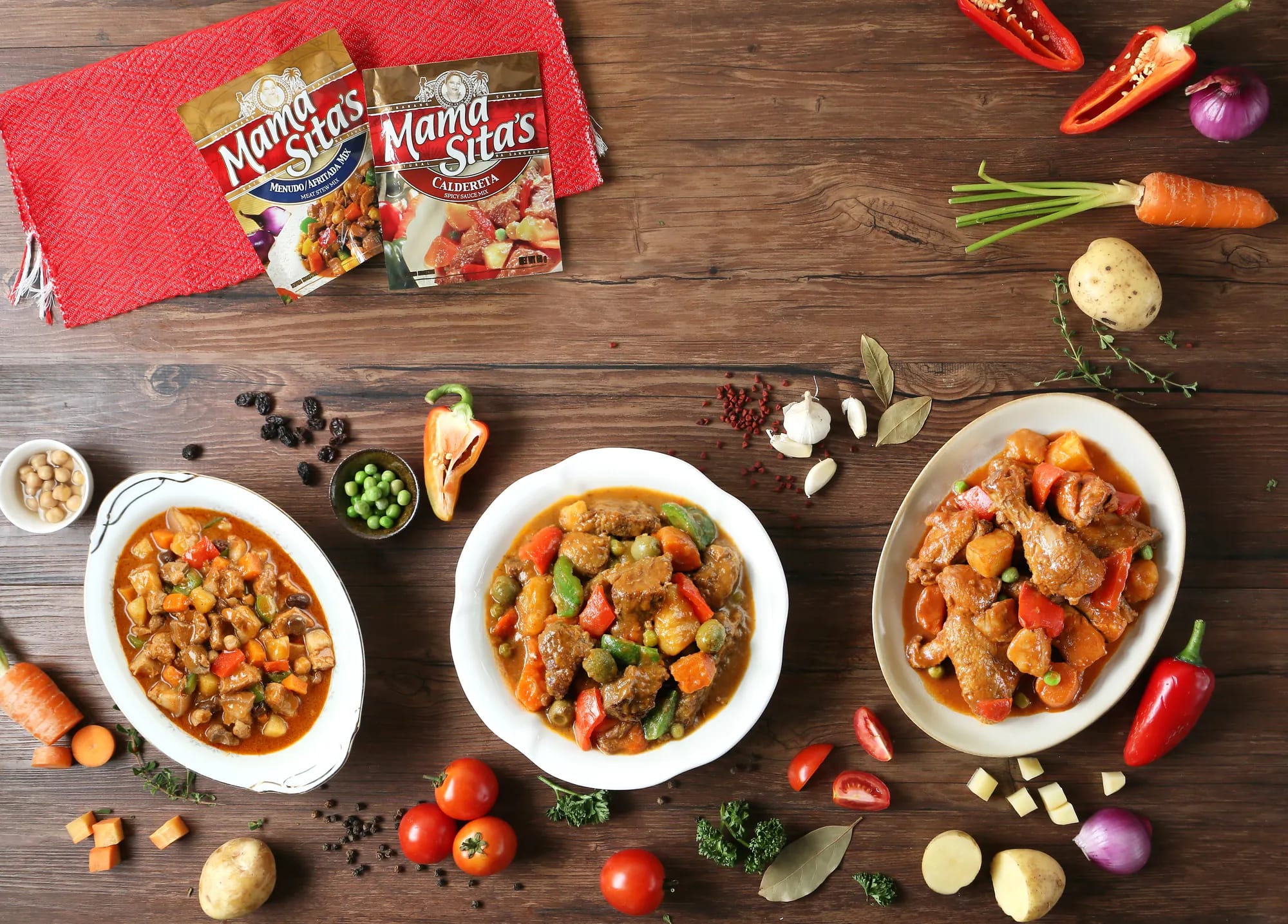 Three white oval platters of pork and chicken stew with potato, carrot and bell pepper, photographed from above on dark wood beside two Mama Sita's seasoning sachets on a red woven mat, with raw vegetables laid around them