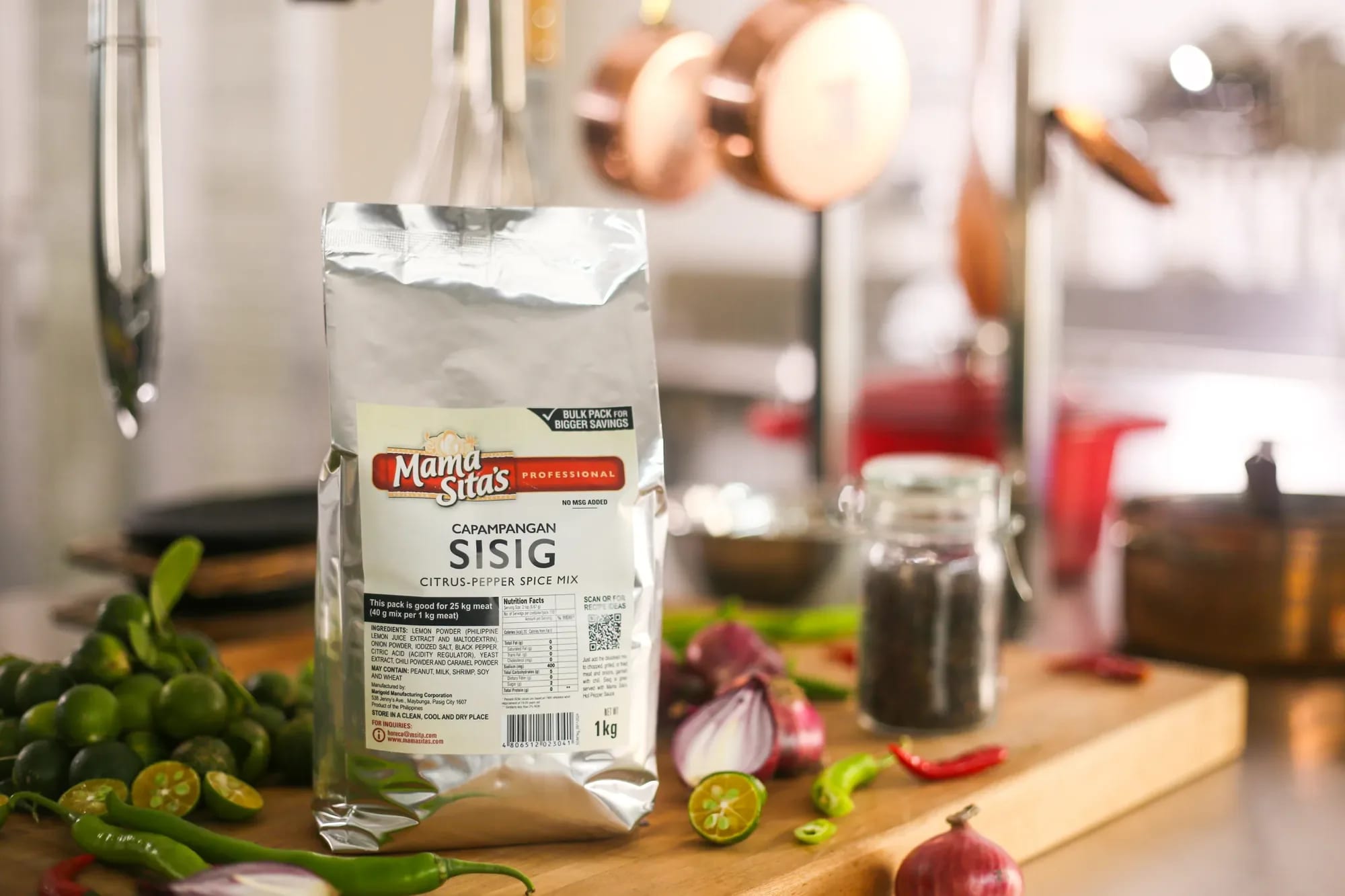 A bag of Mama Sita's Sisig Mix standing on a kitchen counter with fresh chilies and copper pots behind it