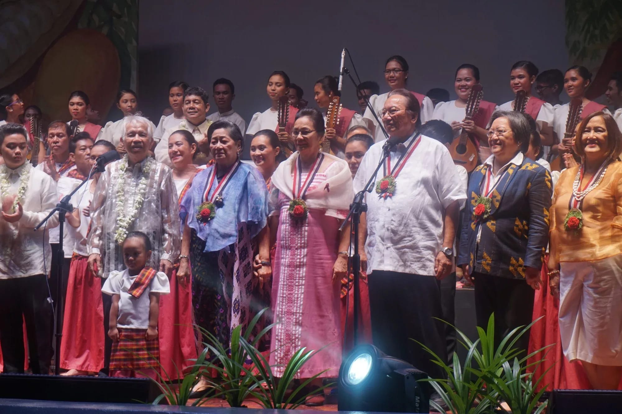 Honorees in Filipiniana standing across a stage under warm light at the Harana centenary tribute, with the Andres Bonifacio Concert Choir and the RTU Tunog Rizalia Rondalla behind them and potted palms at the foot of the stage