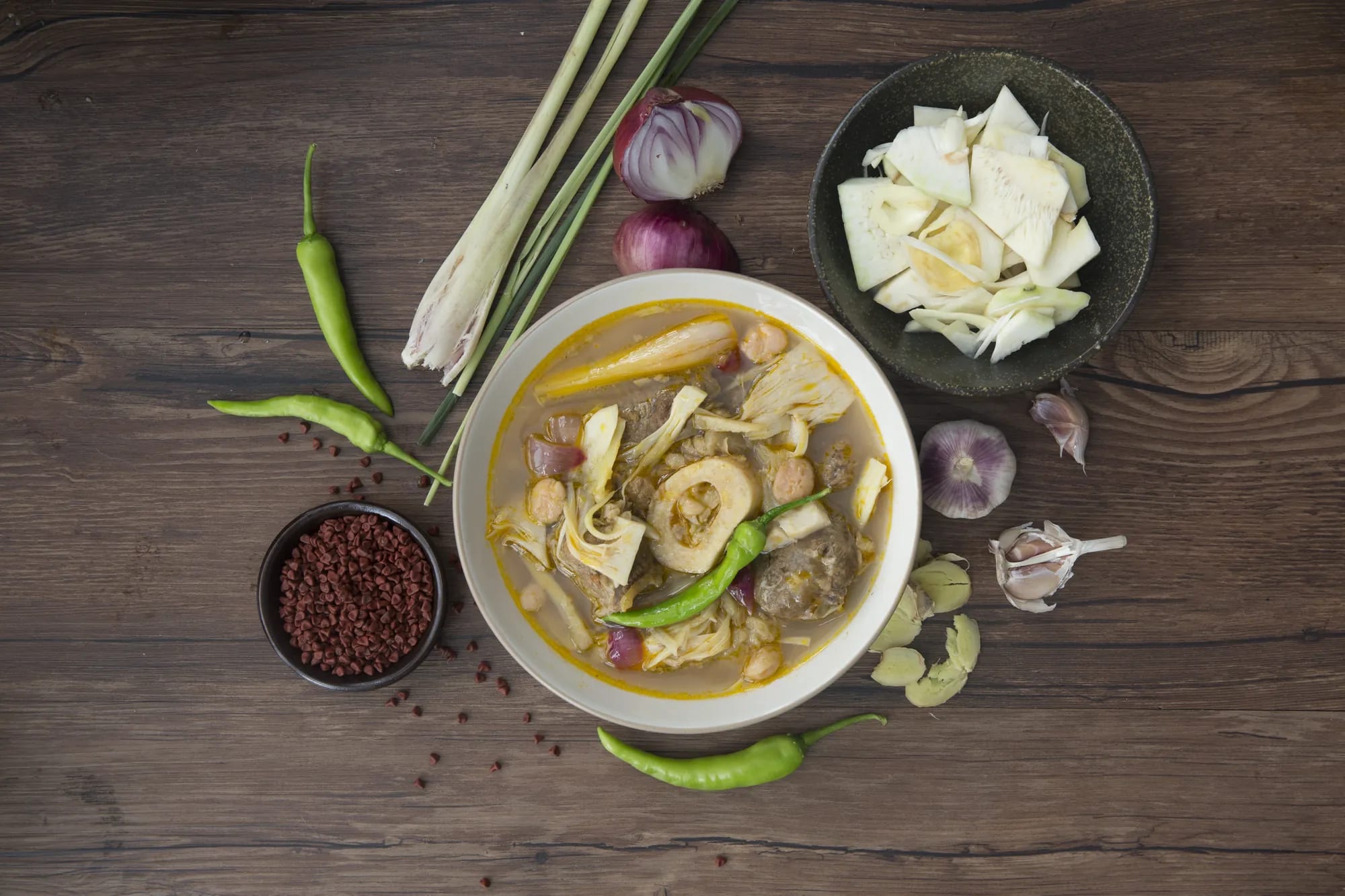 Overhead view of a bowl of beef kansi on a dark walnut table — beef shank, its marrow bone and shaved langka in a lemongrass broth — with lemongrass stalks, halved red onions, green chilies, a bowl of annatto seeds, garlic, sliced batuan and a bowl of shaved young jackfruit around it