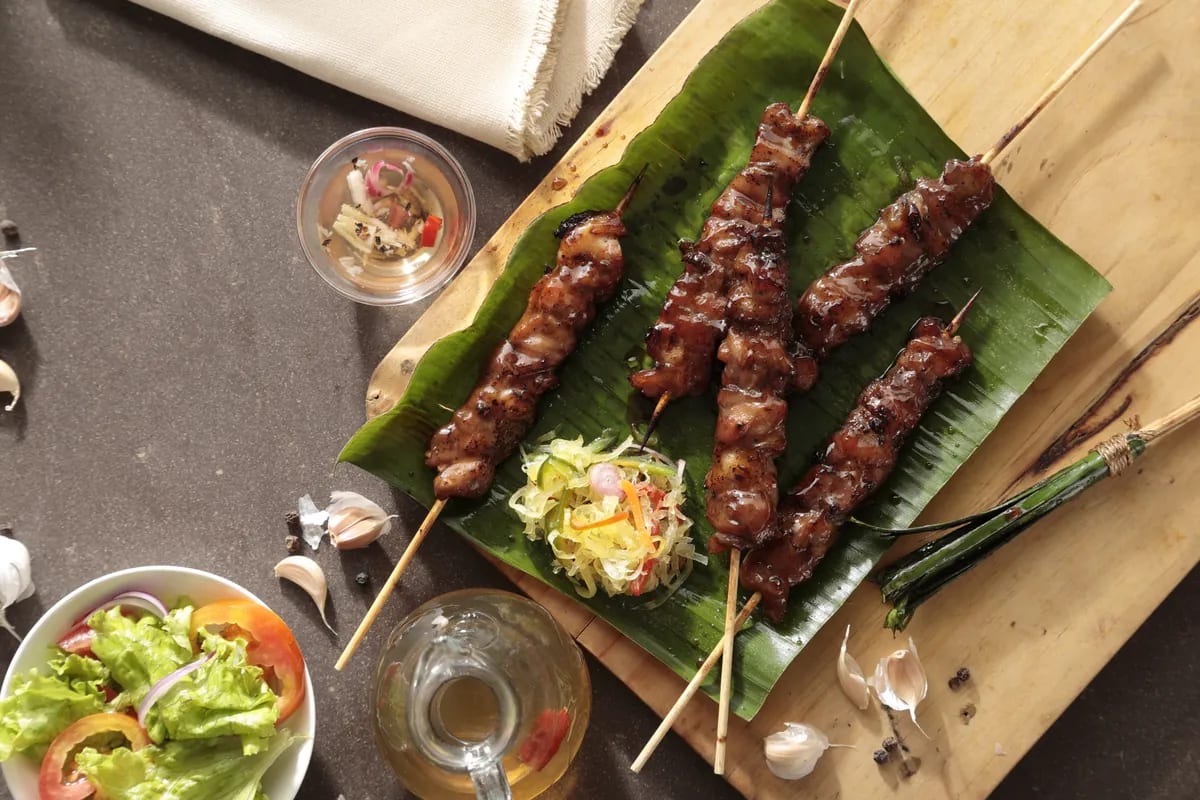 Overhead view of four grilled pork skewers on a banana leaf over a wooden board on dark stone, with a mound of atsara, a glass of atsara in its brine, a cruet of spiced vinegar, a bowl of green salad with tomato and red onion, garlic cloves, peppercorns and a tied lemongrass basting brush around them