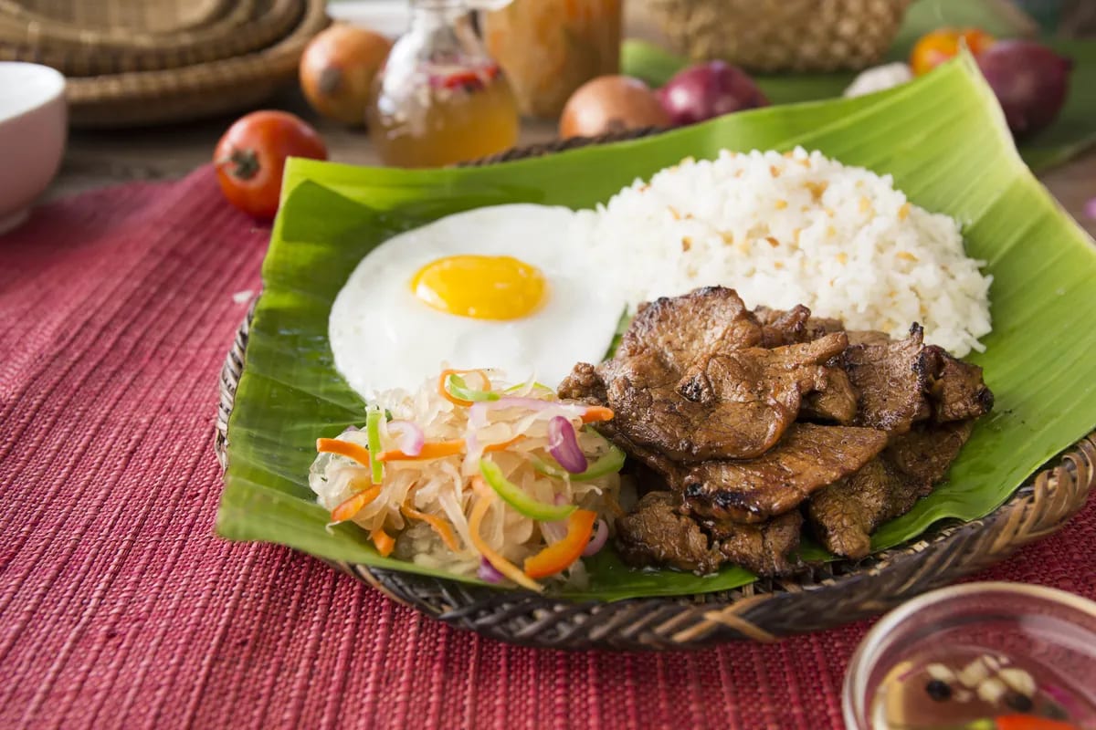 Beef tapa in a woven bilao lined with banana leaf — garlic rice, a fried egg and atsara beside it — on a red woven runner, with a cruet of spiced vinegar, tomatoes and onions soft behind