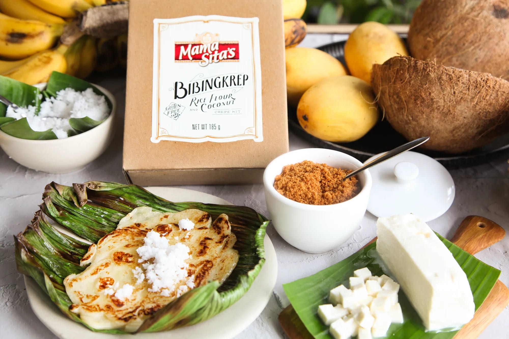 The Mama Sita’s Bibingkrep crepe kit box standing on a white table beside a plate of crepes on banana leaf, with grated coconut, muscovado sugar, kesong puti, mangoes, a coconut and bananas around it
