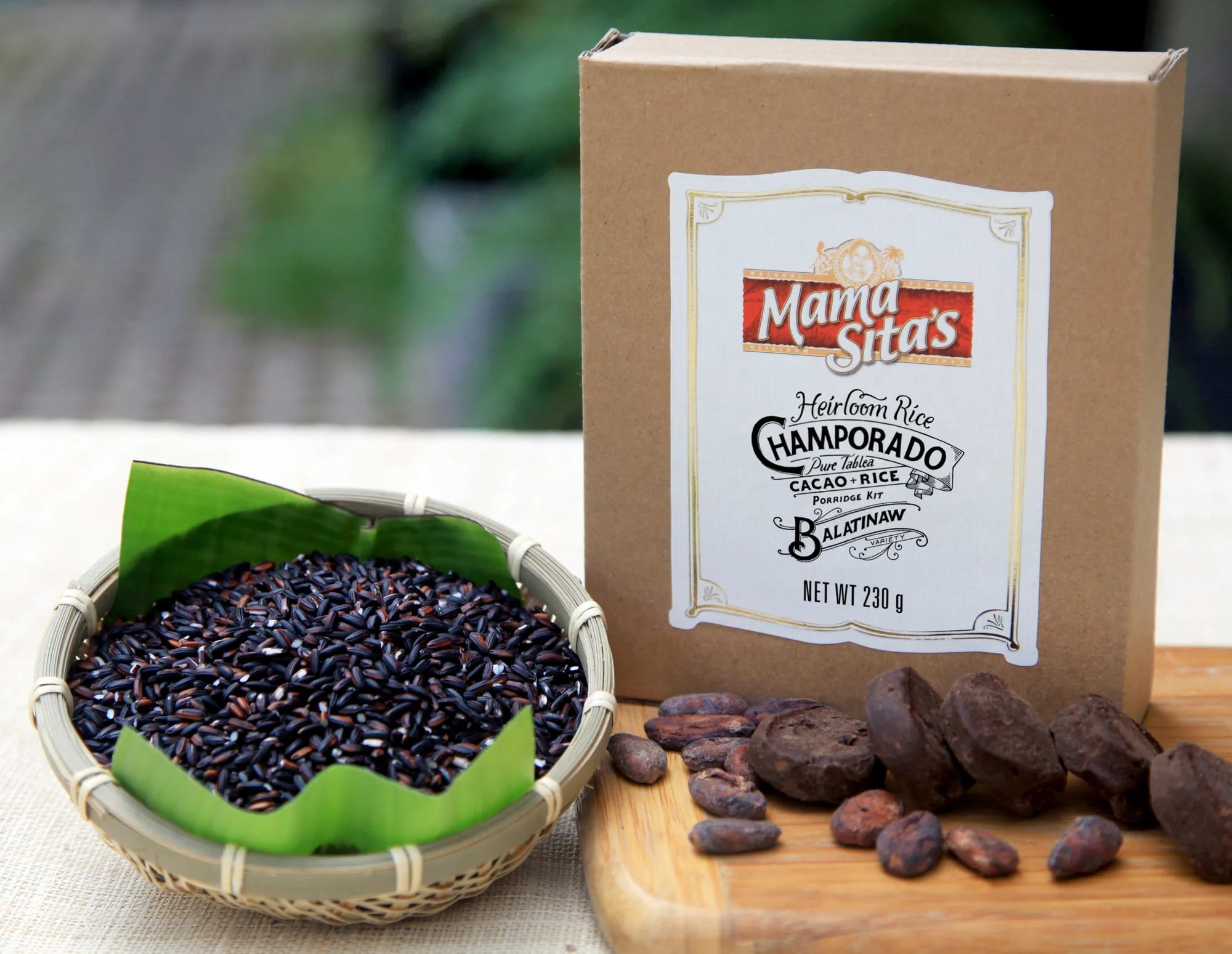 The Mama Sita’s Heirloom Rice Champorado Balatinaw box standing on a table beside a basket of black Balatinaw rice on a banana leaf, with cacao beans and cacao tablea on a board and a garden soft behind