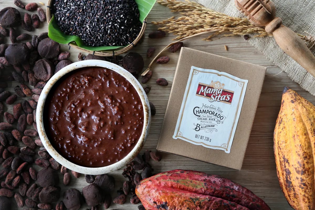 Overhead view of a white bowl of champorado beside the Mama Sita’s Heirloom Rice Champorado Balatinaw box, with cacao beans, cacao tablea, cacao pods, a basket of black rice and rice stalks on burlap