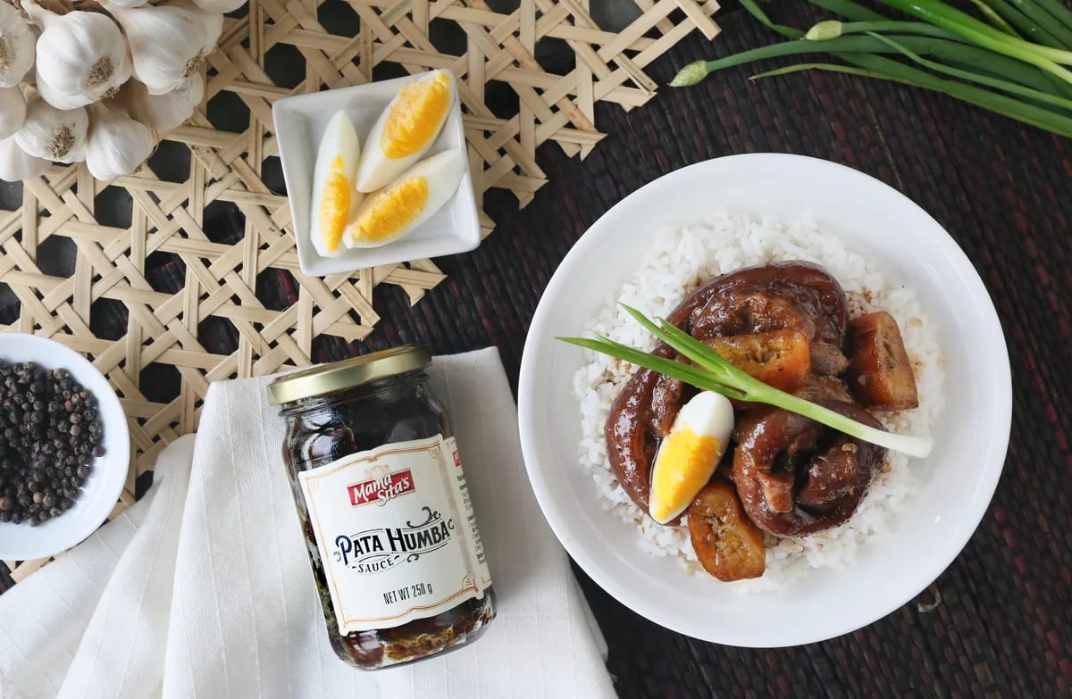A plate of pata humba over rice on a woven placemat, with the Mama Sita’s Pata Humba Sauce jar standing on a folded napkin beside it