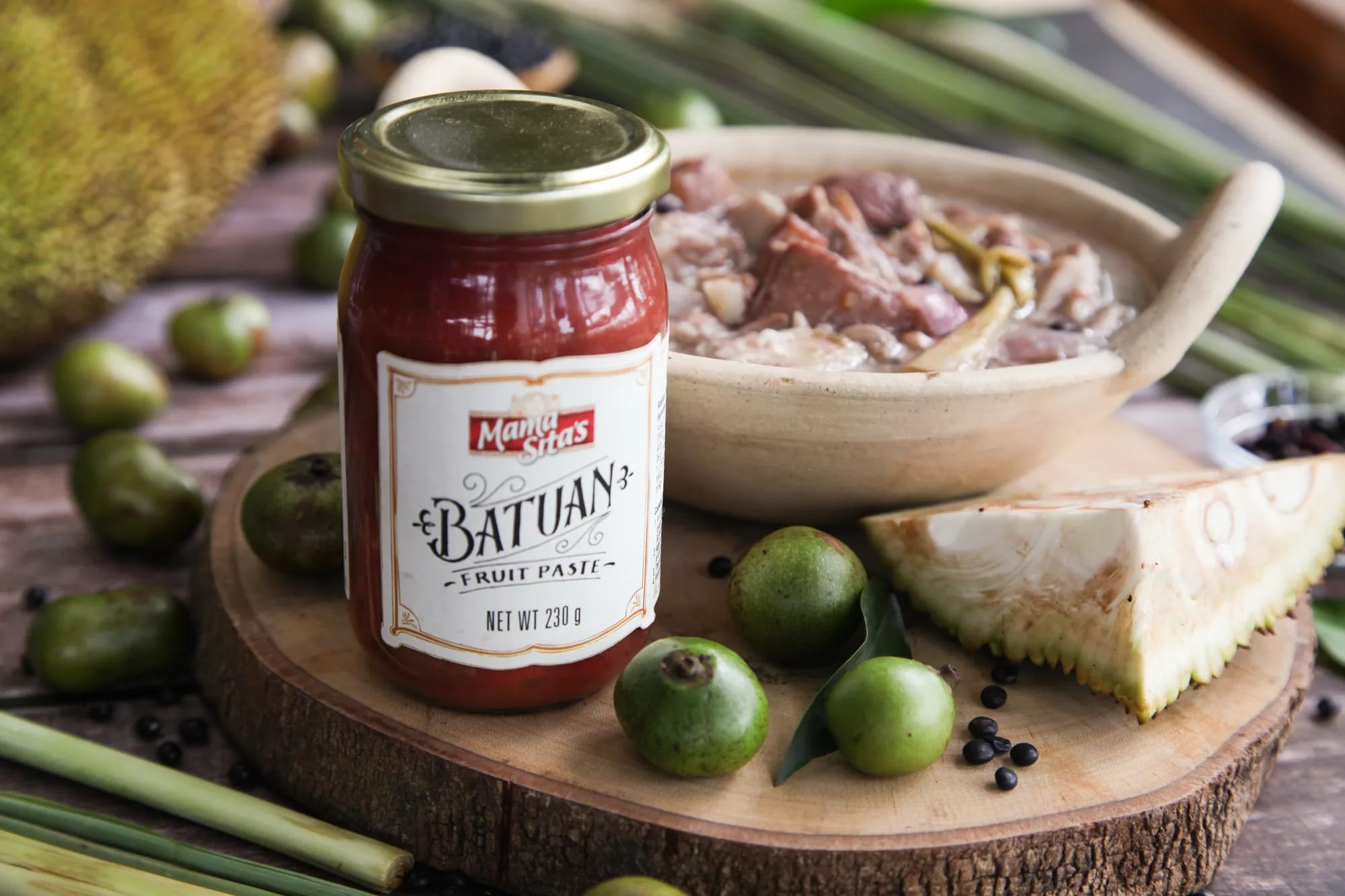 The Mama Sita’s Batuan Fruit Paste jar standing on a round wooden board beside a clay bowl of KBL stew, with batuan fruits, a wedge of jackfruit, lemongrass and peppercorns around it