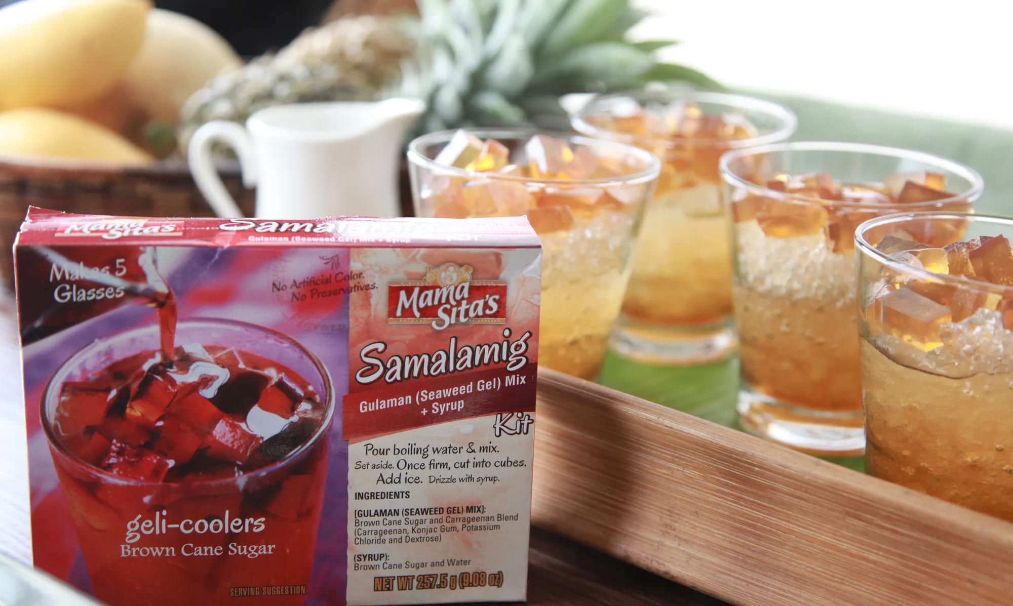 The Mama Sita’s Samalamig Brown Cane Sugar kit box standing on a dark wood counter beside a tray of four tall glasses of gulaman and sago, with a pitcher and mangoes soft behind