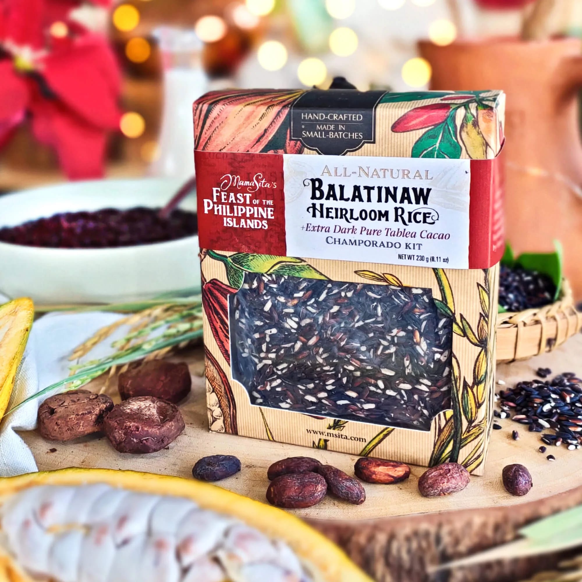 The ribboned Mama Sita’s Feast of the Philippine Islands Balatinaw Heirloom Rice Champorado gift box on a wooden board with cacao pods, cacao tablea, rice stalks and a bowl of champorado, poinsettia and string lights soft behind