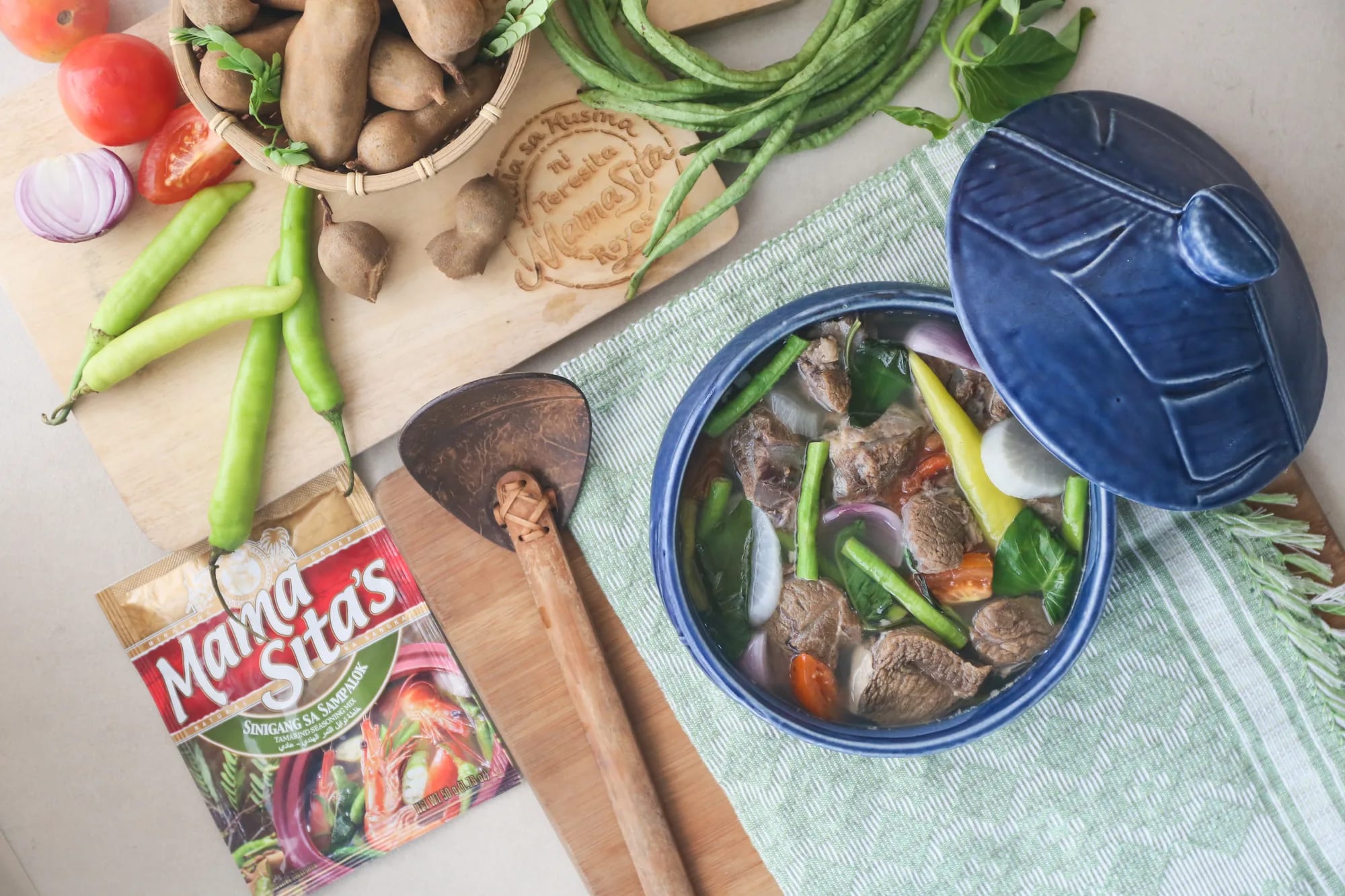 Overhead view of a blue ceramic pot of beef sinigang on a green runner beside a cutting board holding a basket of tamarind pods, tomatoes, red onion, siling haba and a coconut-shell ladle, with the Mama Sita’s Sinigang sa Sampalok Tamarind Seasoning Mix sachet standing at the corner