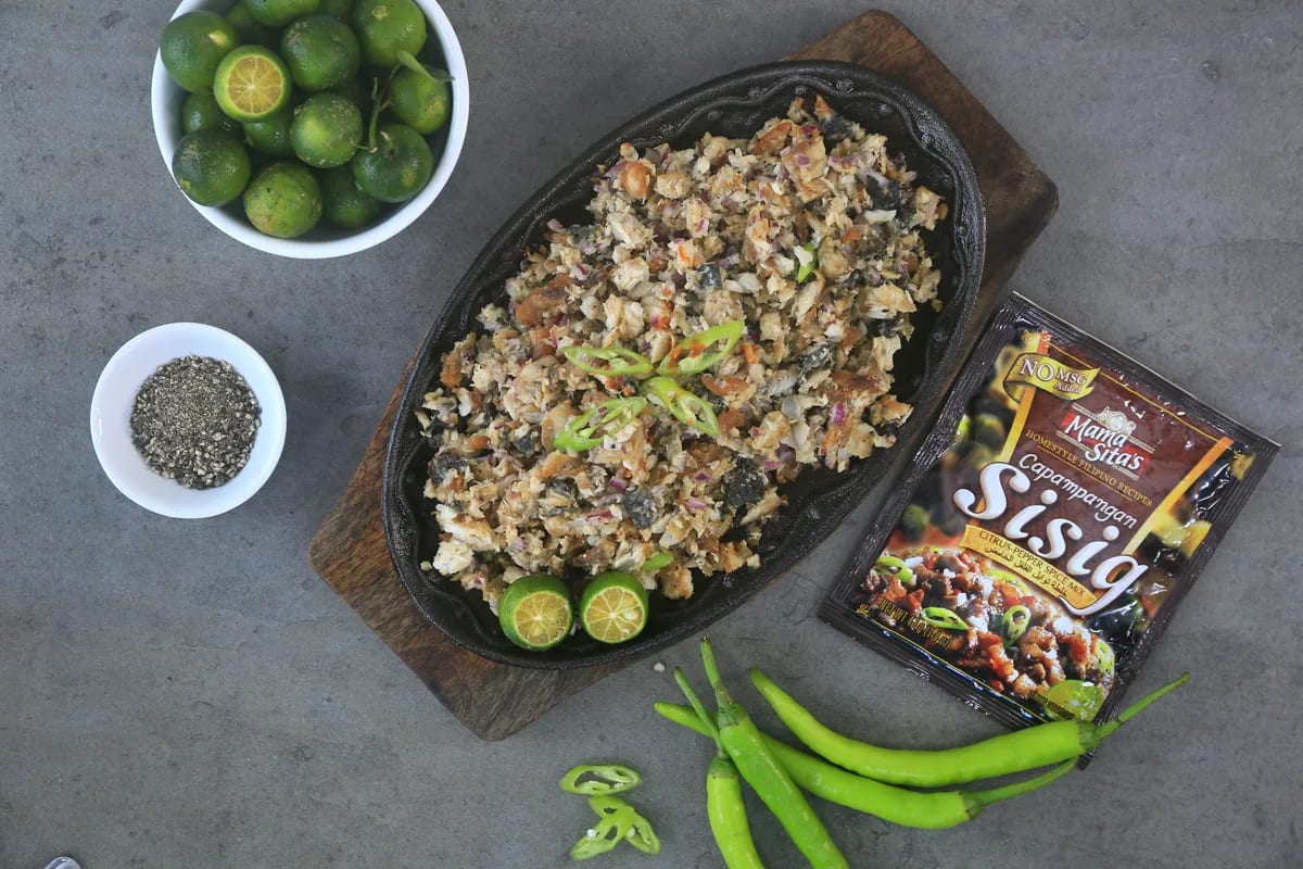 A sizzling plate of bangus sisig on a wooden board beside the Mama Sita’s Capampangan Sisig Mix sachet, with bowls of calamansi and ground pepper and a scatter of green chilies