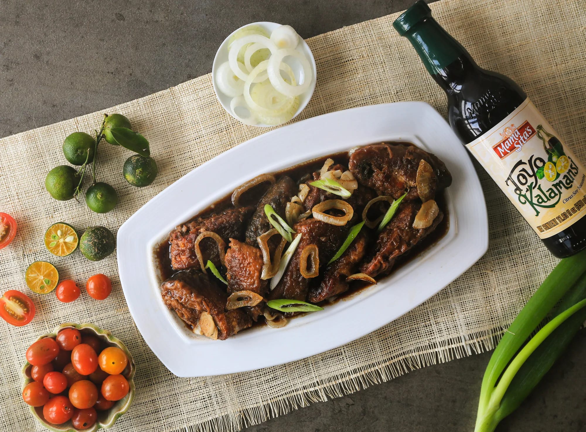 Overhead view of a platter of bistek na bangus with onion rings on a woven placemat, with the Mama Sita’s Toyo’t Kalamansi bottle standing beside calamansi, cherry tomatoes and spring onions