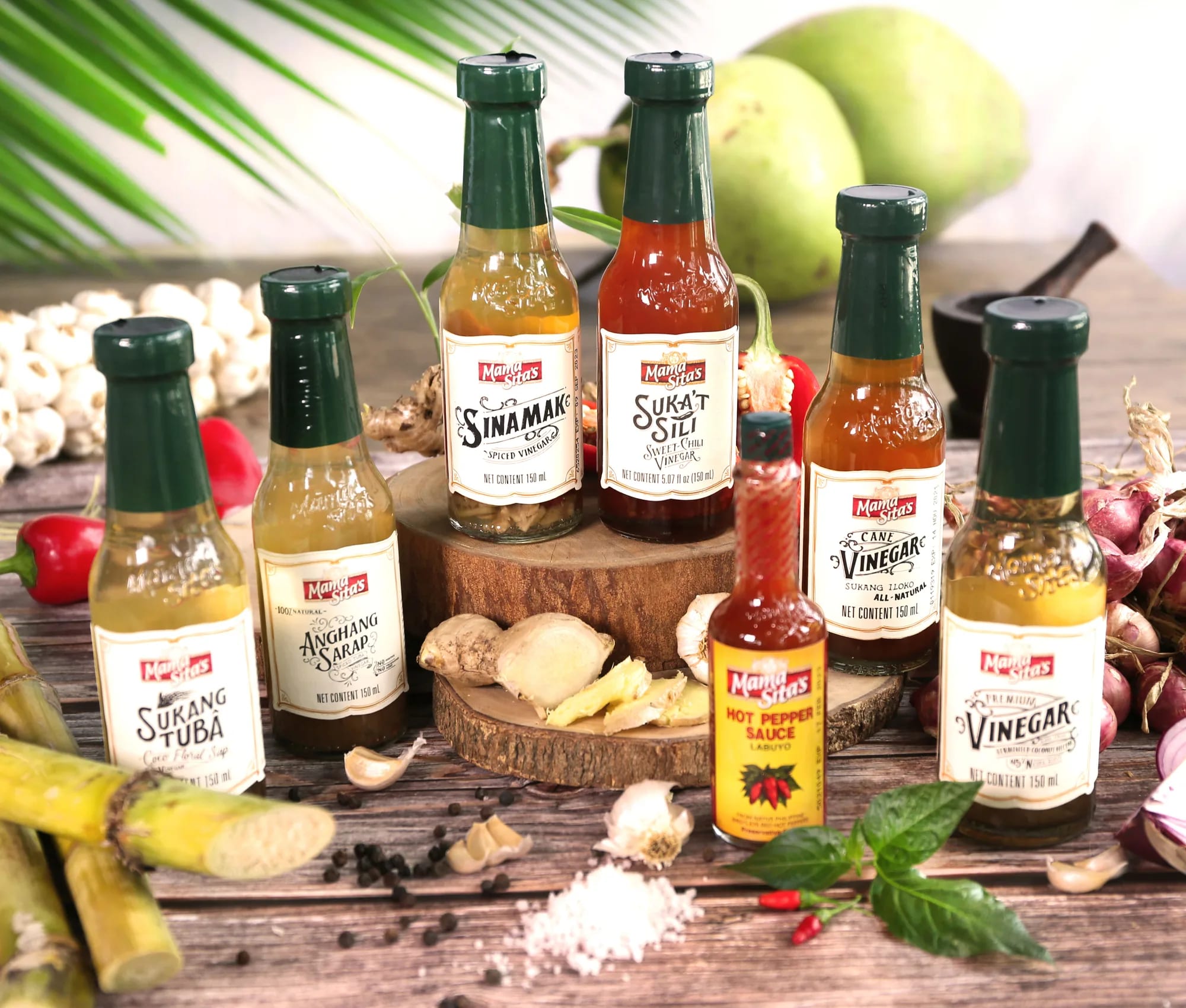 Six Mama Sita’s vinegars standing on a wooden table — Sukang Tuba, Anghang Sarap, Sinamak, Suka’t Sili, Cane Vinegar and Premium Vinegar — with a Hot Pepper Sauce bottle in front and garlic, ginger, red onions, sugarcane and green coconuts around them