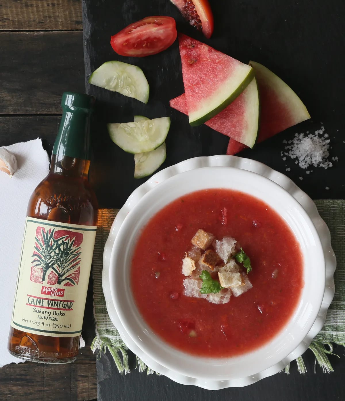The Mama Sita’s Sukang Iloko bottle standing on dark slate beside a bowl of watermelon gazpacho, with tomato, cucumber and watermelon slices around it