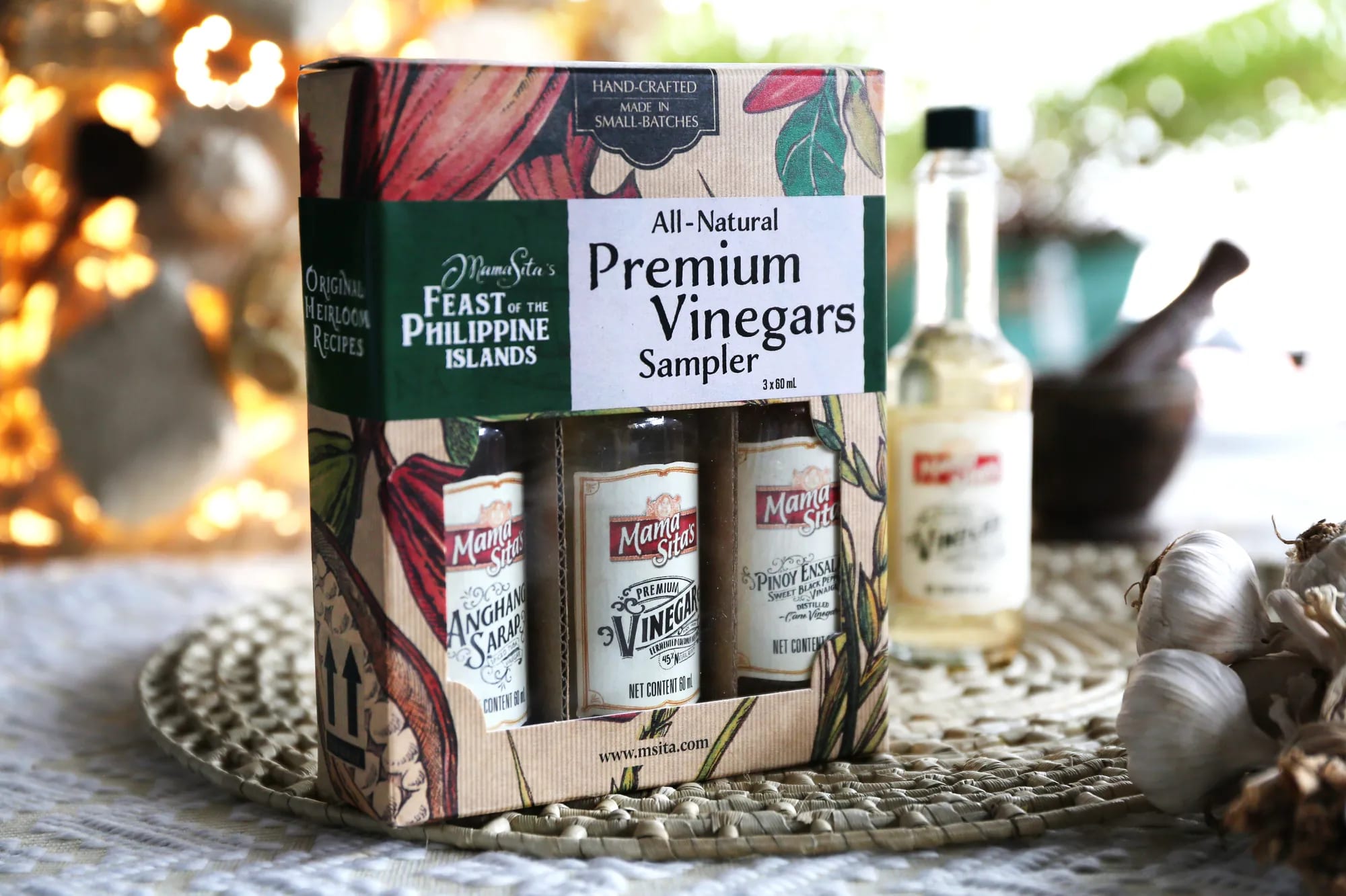 The Mama Sita’s Premium Vinegars Sampler gift box standing on a woven placemat, three small bottles visible through its window, with garlic and out-of-focus string lights behind it