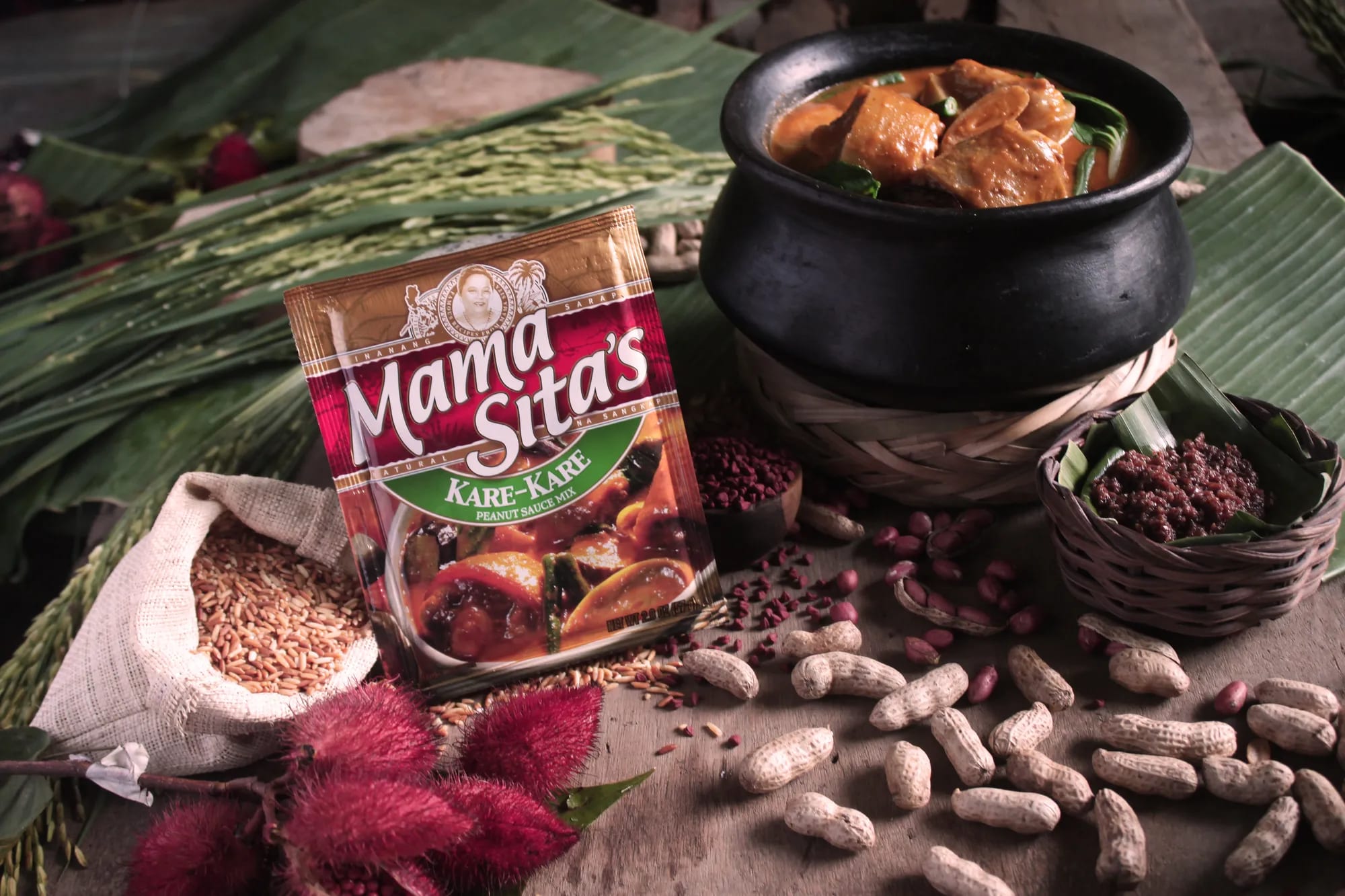 A pouch of Mama Sita’s Kare-Kare Mix standing against a sack of brown rice, with a black clay pot of kare-kare, peanuts and annatto seeds around it
