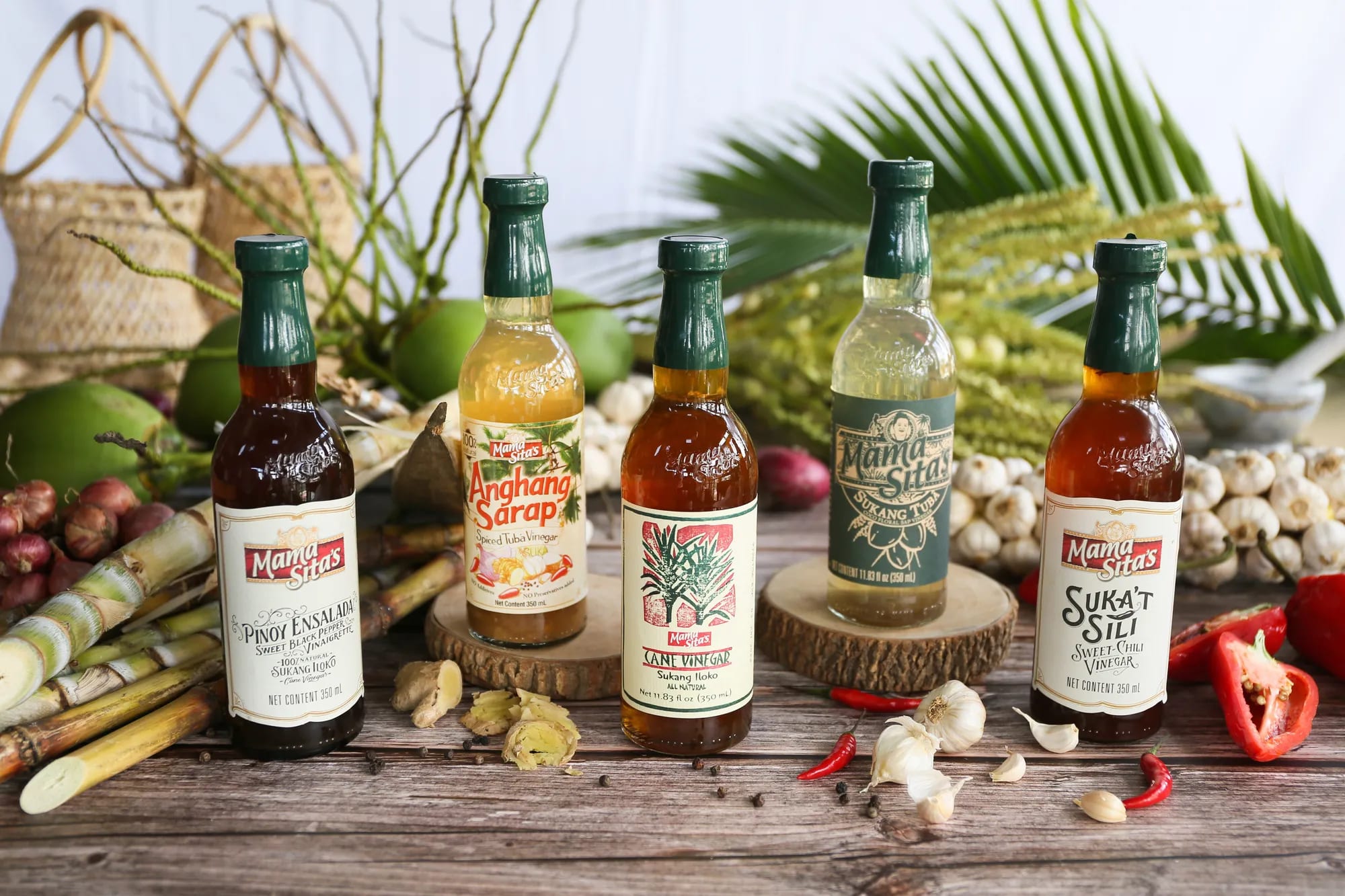 Five Mama Sita’s vinegars standing together on a rustic wooden table — Pinoy Ensalada, Anghang Sarap, cane vinegar, Sukang Tuba and Suka’t Sili — among sugarcane, garlic, chilies, green coconuts and buri baskets