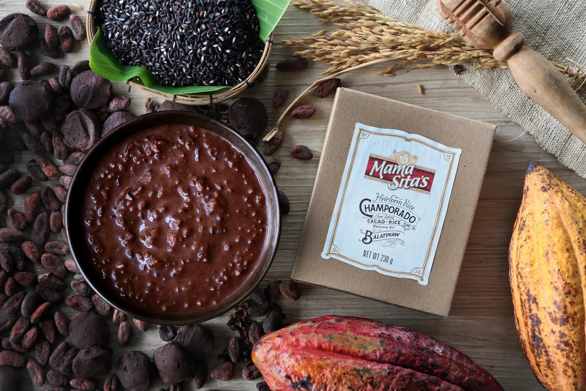 A bowl of champorado beside the Mama Sita’s Heirloom Rice Champorado box, with cacao pods, cacao tablea and black Balatinaw rice around them