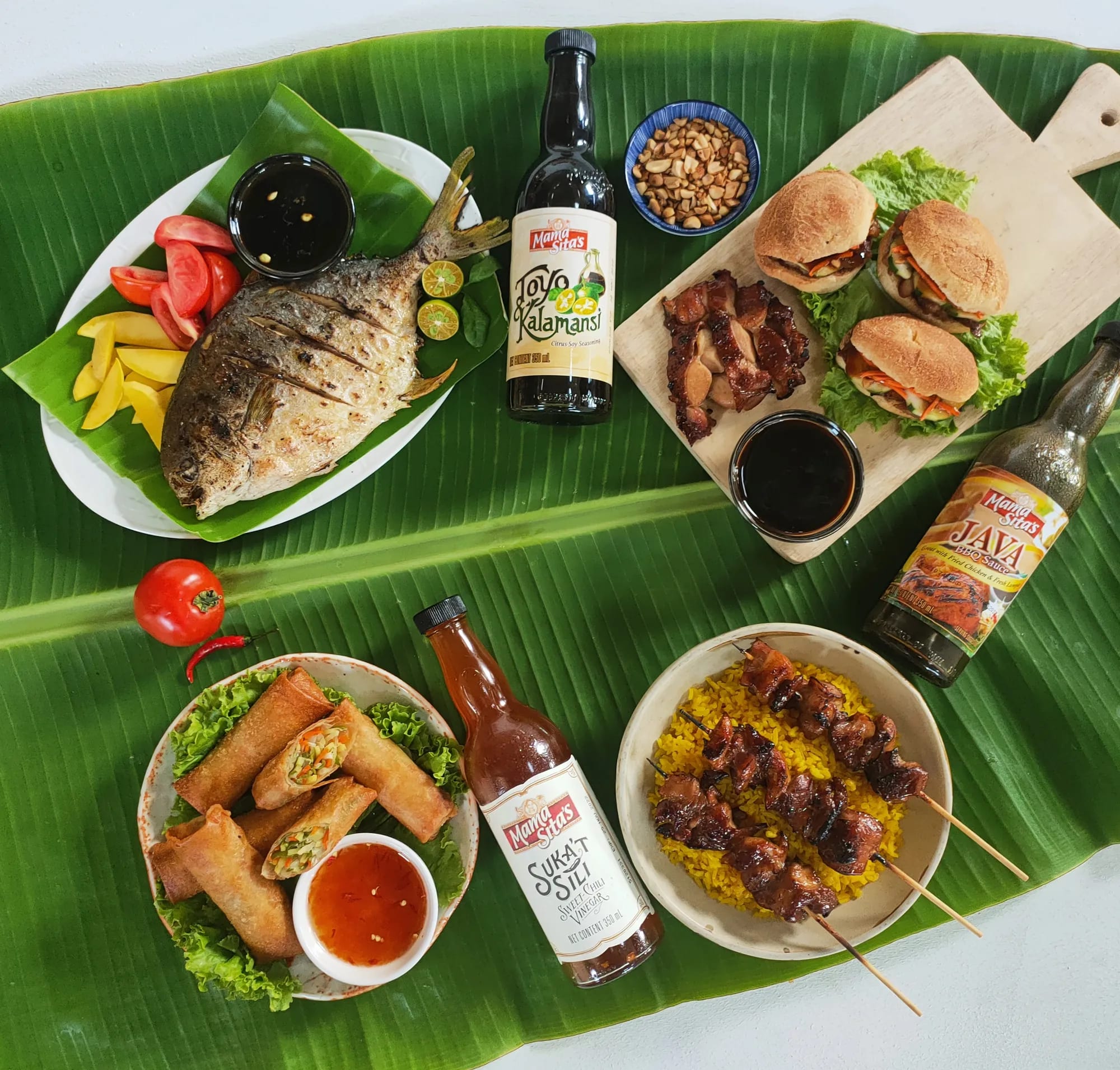 An overhead banana-leaf spread — grilled fish with sawsawan, barbecue pork sliders on a board, lumpia with sweet chili dip, and skewers over yellow rice — with Mama Sita’s Toyo’t Kalamansi, Java Barbecue Sauce and Suka’t Sili standing among the dishes